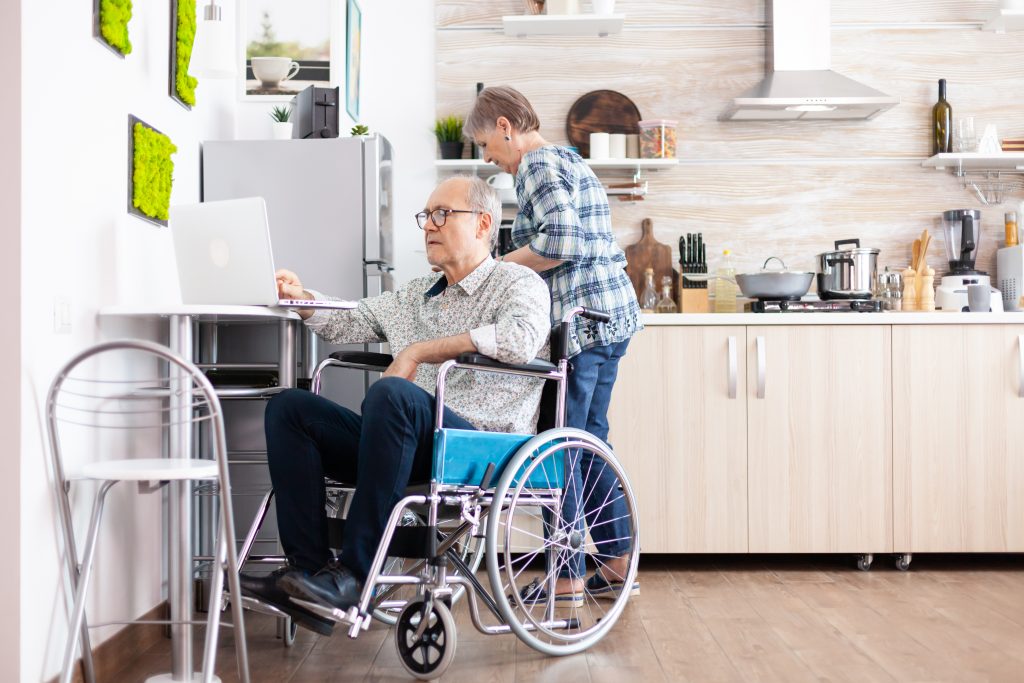 Elderly man in a wheelchair typing in a laptop, working from home while wife is cooking breakfast handicapped businessman entrepreneur paralyzed retired elderly male