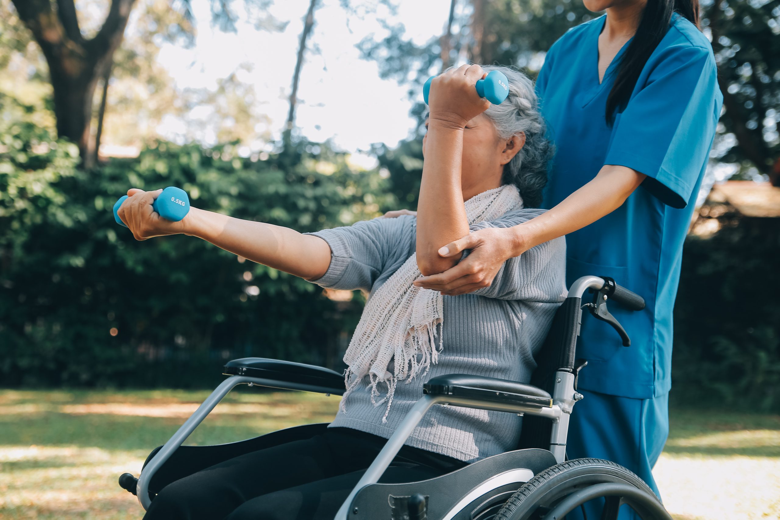 Una joven enfermera o fisioterapeuta, vestida con ropa de trabajo, ayuda a una anciana jubilada y alegre a hacer ejercicios de fitness con mancuernas ligeras en casa. Concepto de fisioterapia para personas mayores